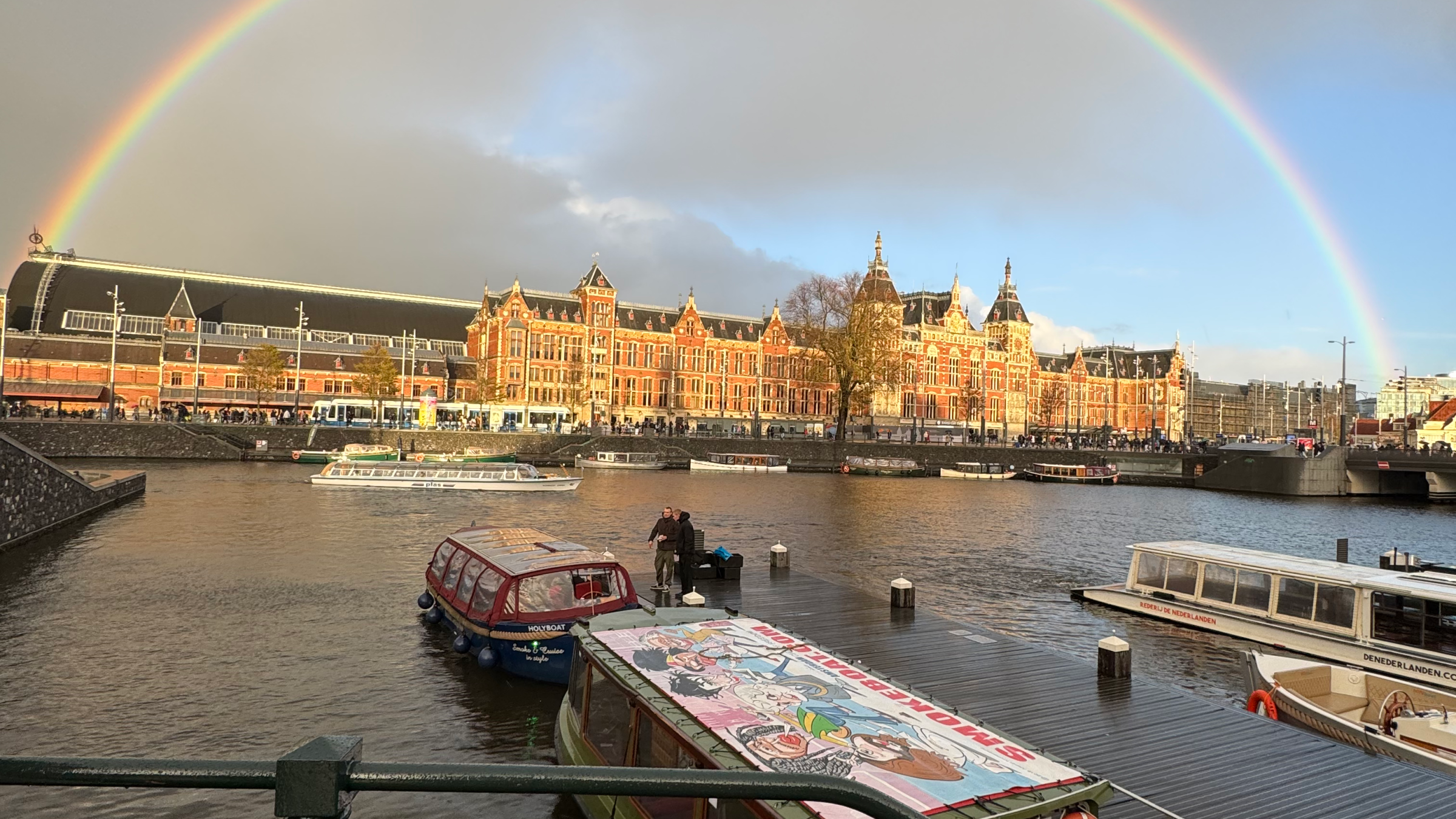 Rainbow over historic buildings and boats on a canal under a cloudy sky.