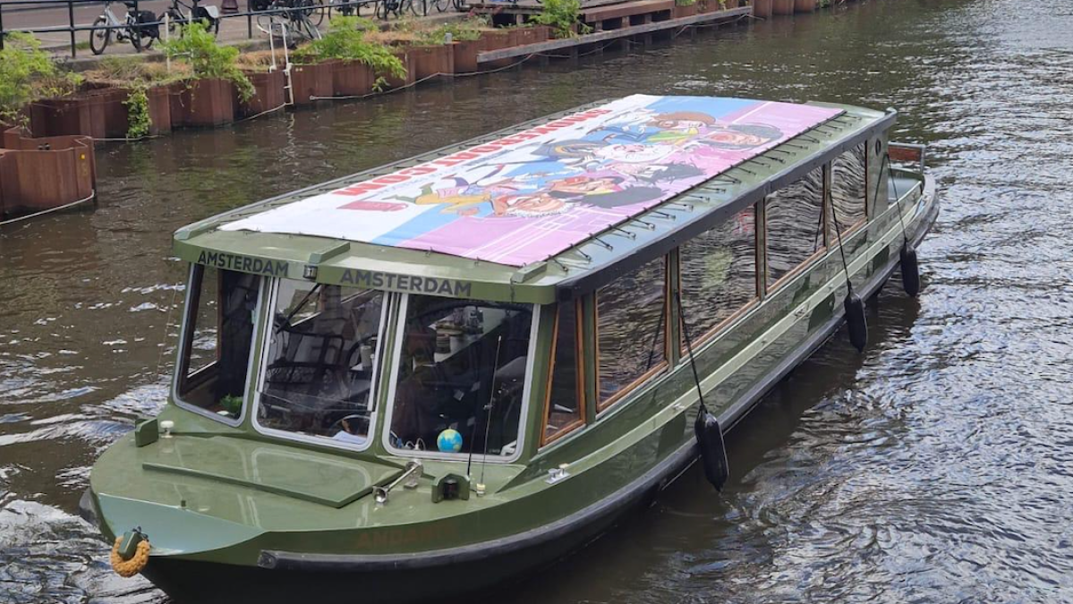 Green canal boat on a waterway flanked by brick buildings and trees.