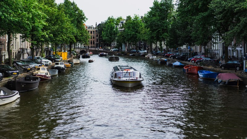 Boat on a tree-lined canal with parked boats and buildings in the background.